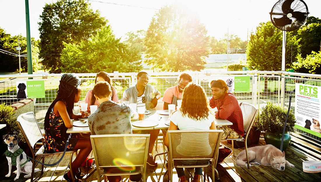 A group of people sit around a table on an outdoor patio. The space is fenced in and has signs about pet owner responsibility on the fencing. Dogs are tethered to the fence and the table and a fan aims cool air down toward the dogs. There is a water bowl on the patio for dogs too.
