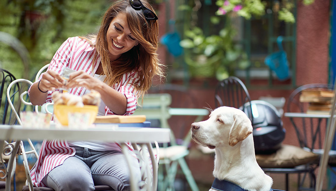 A woman sits at a cafe table with a large tan dog sitting by her side.