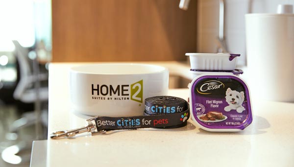 A water bowl with the Home2 Suites by Hilton logo sits next to a package of CESAR dog food and a dog leash.