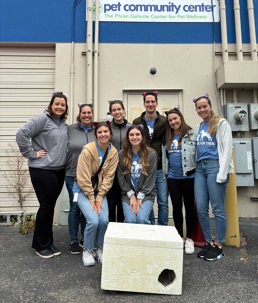 A group stands in front of Pet Community Center with a winter cat shelter in front of them. They're wearing cat-ear headbands with the left ear clipped - a signal that a cat has been part of a trap-neuter-return program.