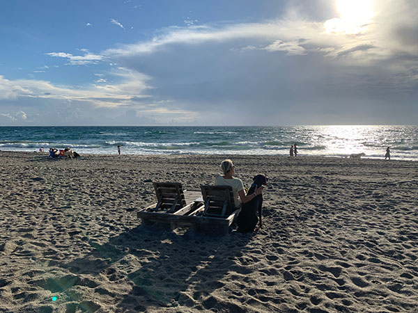 Pet pet parent and their dog sit on the beach enjoying the view at Spanish River Dog Beach in Boca Raton