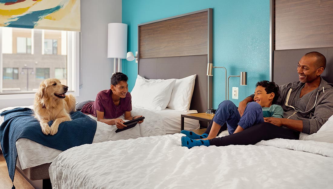 Une famille est assise sur des lits dans un hôtel Tru by Hilton. Ils ont un golden retriever avec eux qui se repose joyeusement.